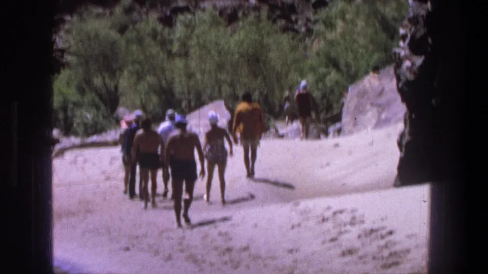 Soft 8mm frame of a 1969 Utah outdoor outing, with adults in swimwear walking away through a sandy rocky area.