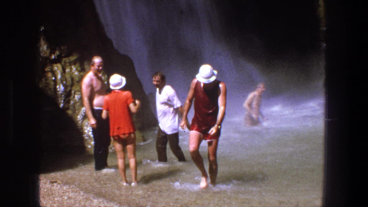 1969 Utah home-movie frame showing adults in period swimwear wading by a misty waterfall and dark rock face.