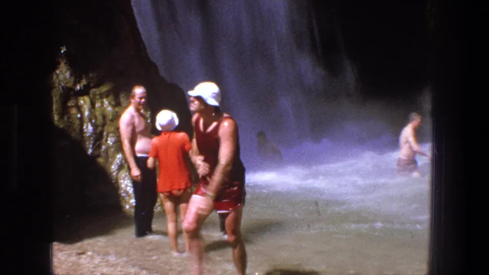 Adults in classic swimwear and sun hats stand and talk in shallow water near a powerful Utah waterfall.