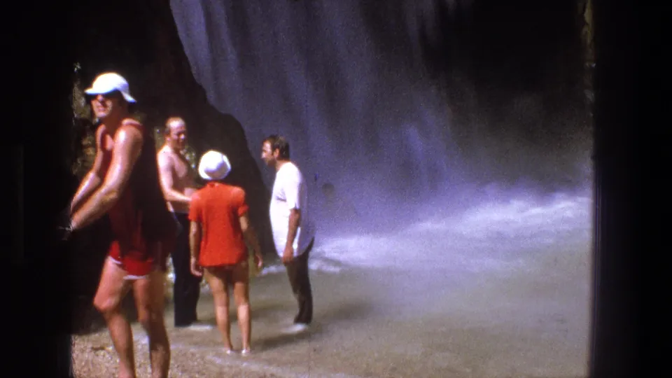 A handheld 8mm frame shows adults shifting positions in shallow water at a rocky waterfall site in Utah.