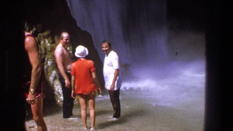 Adults in 1960s swimwear stand close to the camera at a Utah waterfall, with mist and rushing water behind them.