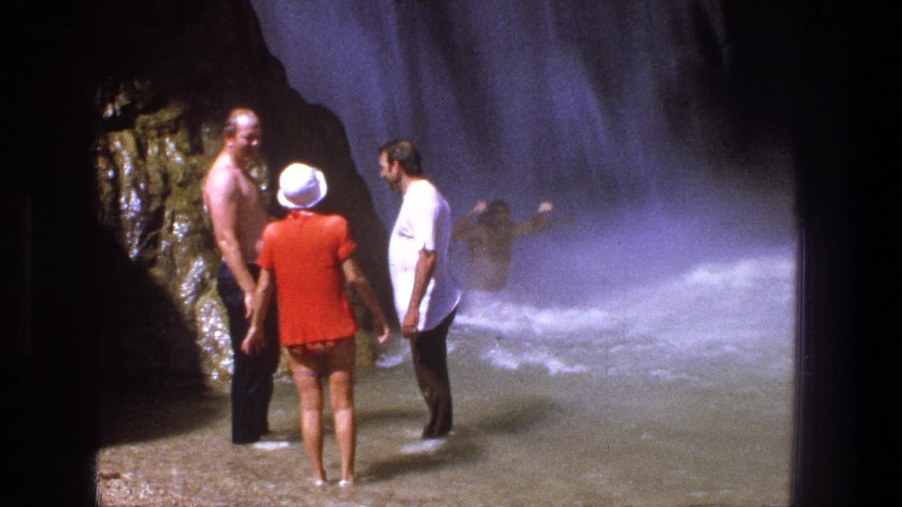 1969 Utah home-movie frame of adults standing in shallow water near a rocky waterfall as mist and falling water fill the background.