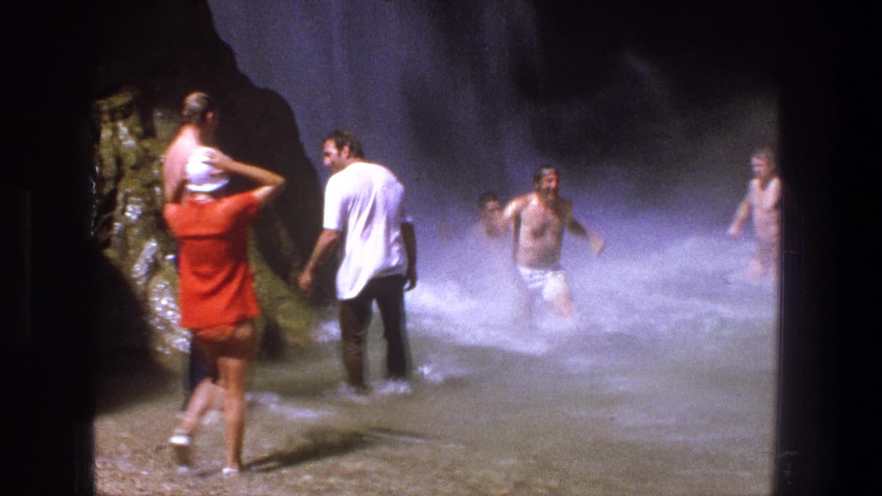 A person in a red shirt carries a child-sized figure or small person on their shoulders while adults wade near the waterfall.