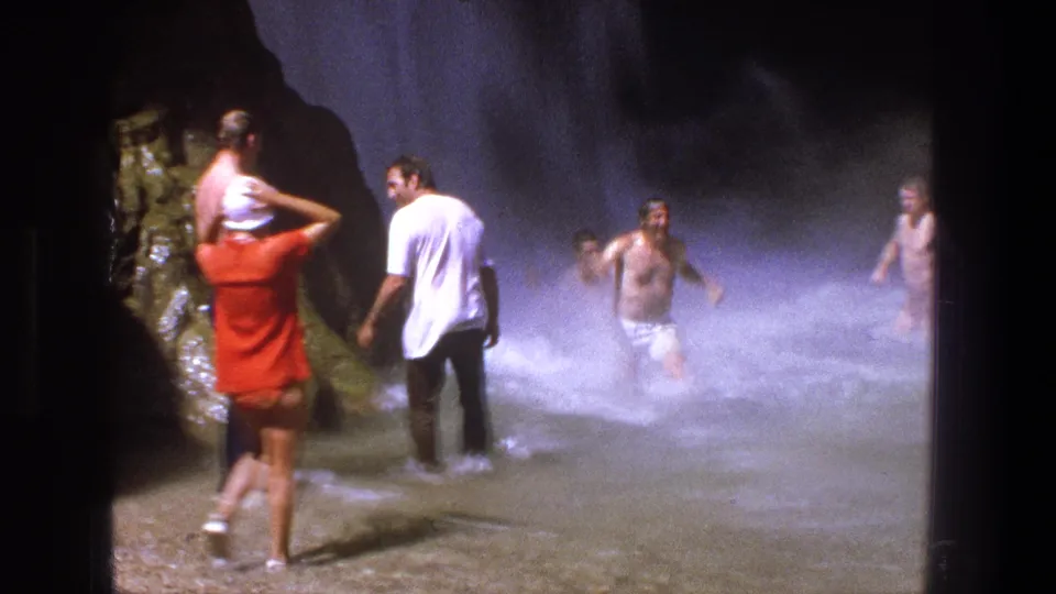 1969 Utah waterfall scene with adults wading through misty water; a person in red appears to carry someone on their shoulders.
