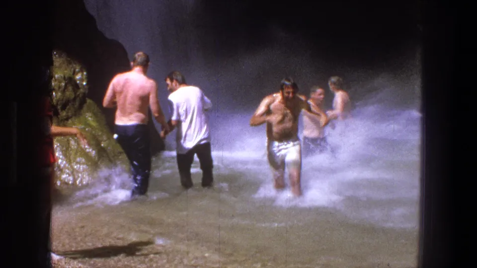 1969 Utah 8mm frame of a wet man in white shorts emerging from the waterfall pool as other adults remain near the spray.
