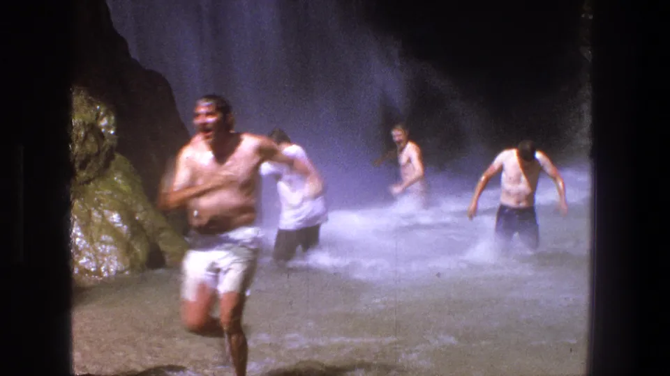 1969 Utah home-movie frame of a shirtless man stumbling out of shallow waterfall water, with mist and other adults behind him.