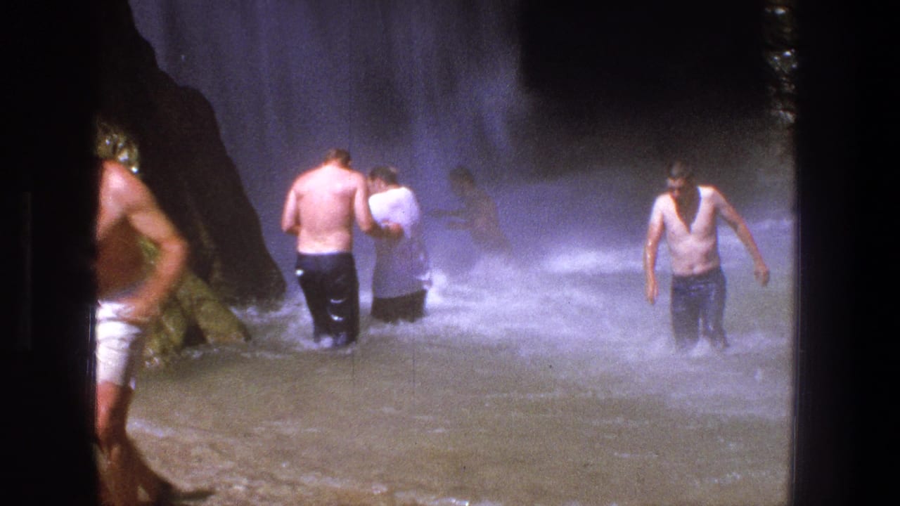 Several men wade in shallow water near dark rocks and waterfall spray.