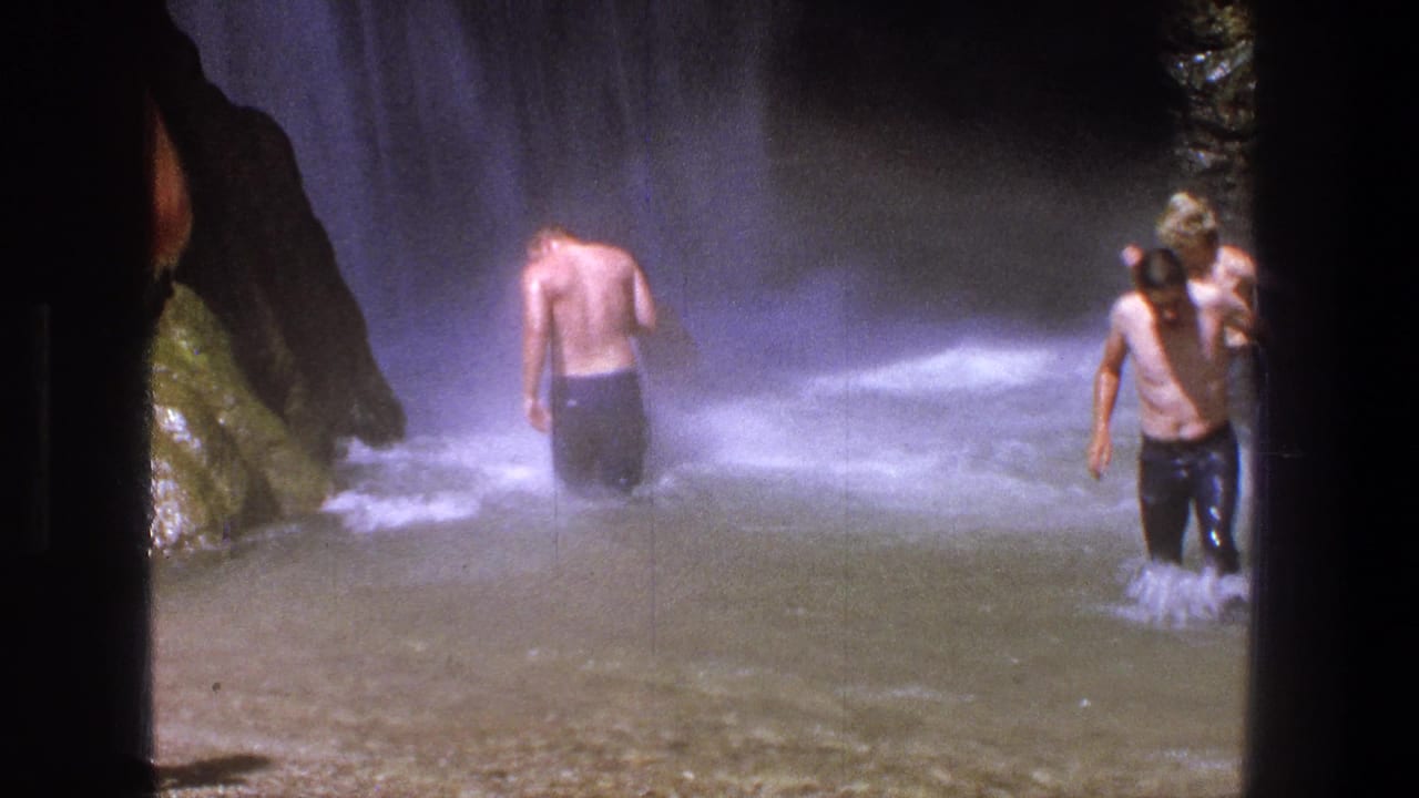 Two shirtless men stand in misty water beside a waterfall and rocks.