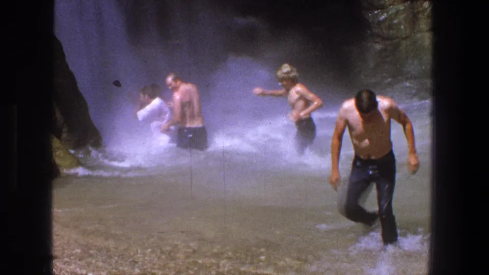 A group of men wades through the waterfall pool, with one man stepping toward camera through spray in 1969 Utah 8mm footage.