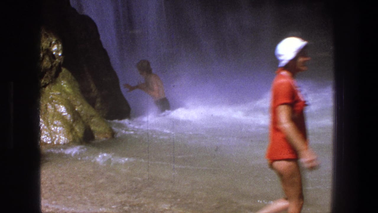 Woman in a red swimsuit walks through shallow water near a strong waterfall and rocks.