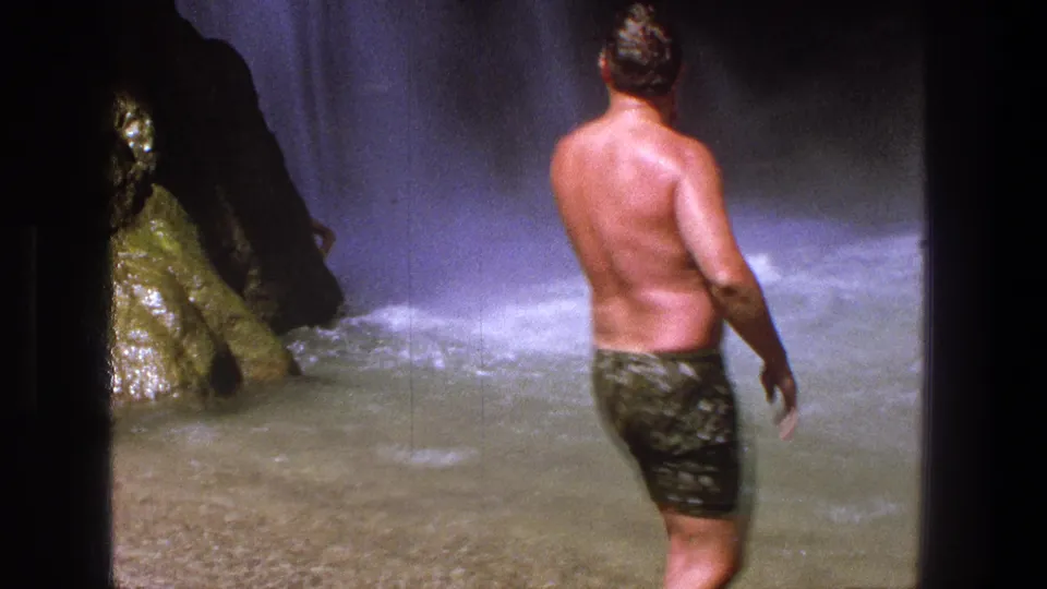 1969 Utah home-movie frame of a shirtless man in patterned trunks watching a forceful waterfall from shallow water.