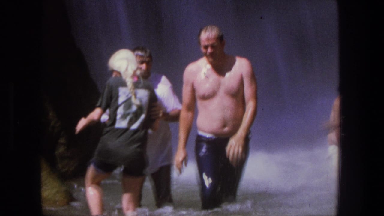 In 1969 Utah home-movie footage, wet adults move through shallow water near a waterfall, including a shirtless man and a woman in dark clothing.