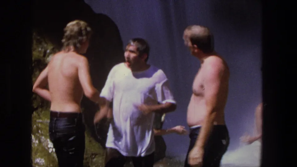 In the final frame, soaked adult men face one another in conversation beside the waterfall, with mist and a rocky edge behind them.
