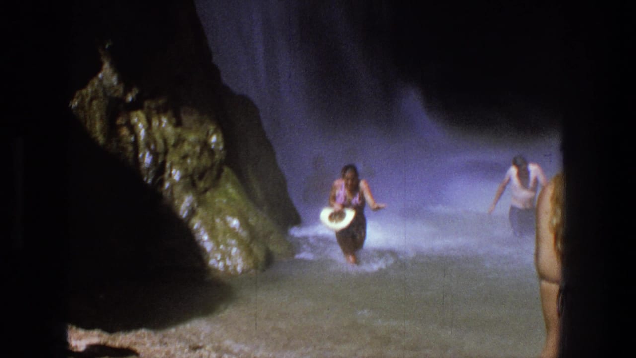 A woman carrying a white hat wades through shallow water between rocks.