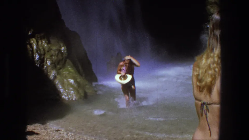 1969 Utah 8mm frame showing a woman carrying a white hat while wading toward the camera through shallow canyon water.