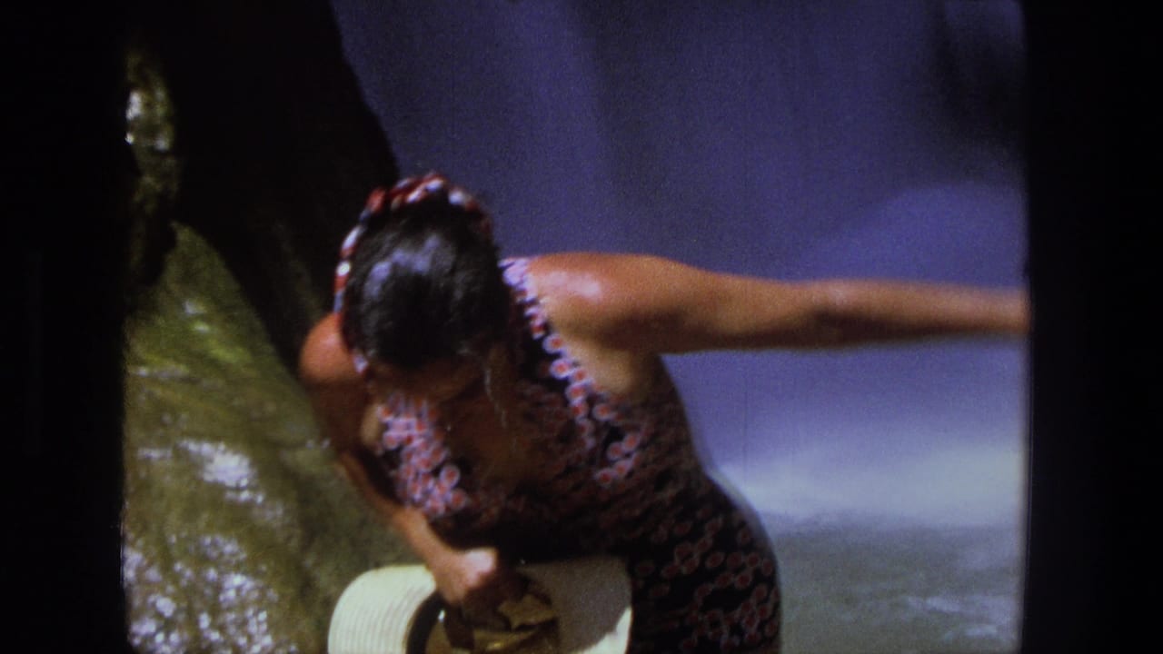 The woman leans out from the waterfall spray, wet hair slicked back and one arm extended beside the rock face.