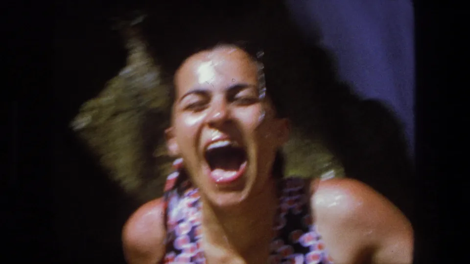 A close 8mm frame shows the drenched woman laughing openly, with waterfall spray and dark rock behind her.