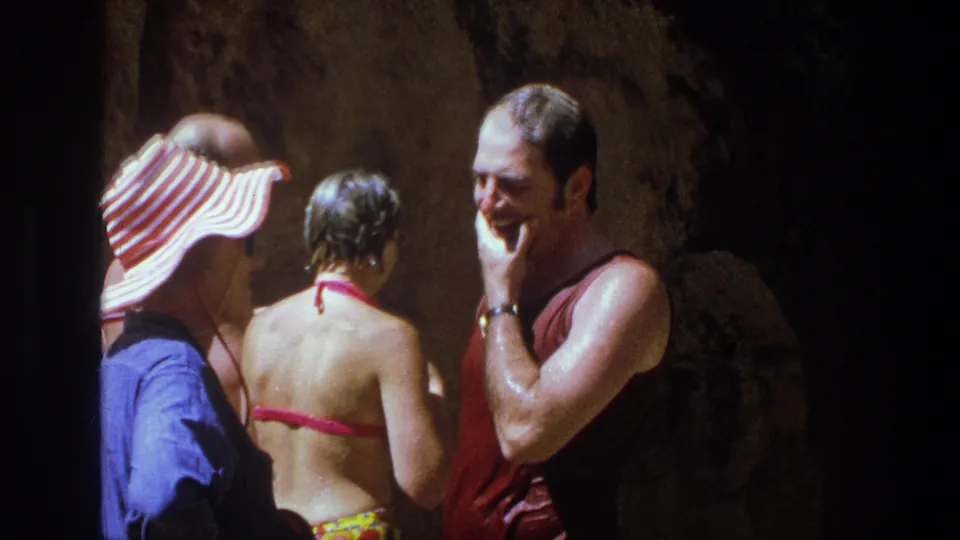 A man in red sleeveless swimwear smiles or laughs beside two other adults in a rocky Utah waterfall setting.