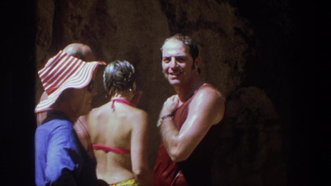 In 1969 Utah home-movie footage, adults in swimwear gather in a dim cave-like setting after swimming, with a man in a red tank top facing camera.