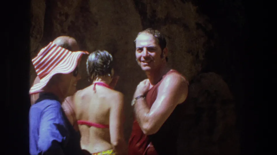 In 1969 Utah home-movie footage, adults in swimwear gather in a dim cave-like setting after swimming, with a man in a red tank top facing camera.