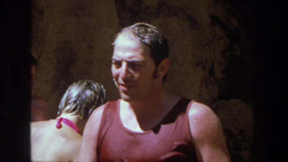 The home-movie frame shows a wet man in a red tank top looking down as a woman in a pink bikini stands behind him in the Utah cave-like setting.