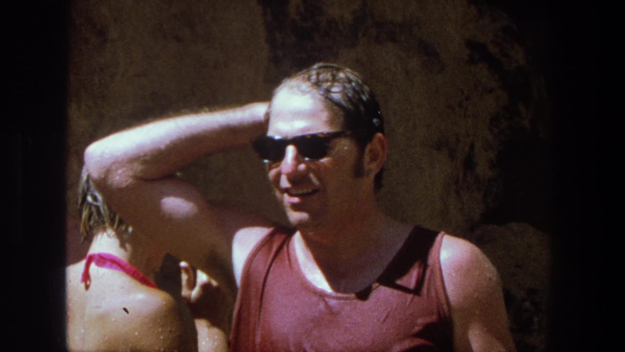 1969 Utah home-movie frame showing a smiling wet man in sunglasses and a woman turned away near a rocky wall.