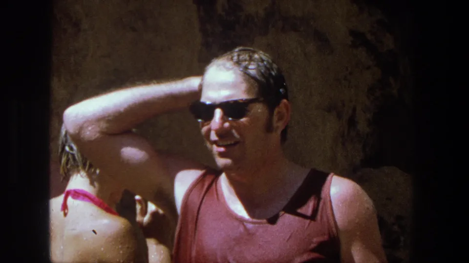 1969 Utah home-movie frame showing a smiling wet man in sunglasses and a woman turned away near a rocky wall.