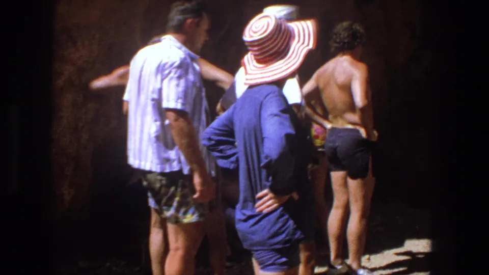 1969 Utah home-movie still with adults in swimwear standing by rough rock, including a woman in a striped hat and blue cover-up.