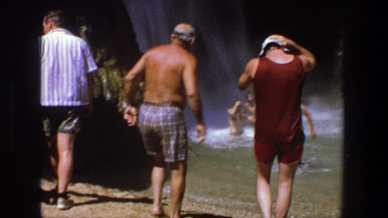 Adult men in swimwear stand by the misty pool, with one man adjusting a white cap in the spray.