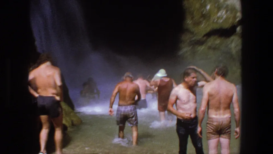 Several adult men in swim trunks gather in the shallow water below a shaded waterfall in Utah.