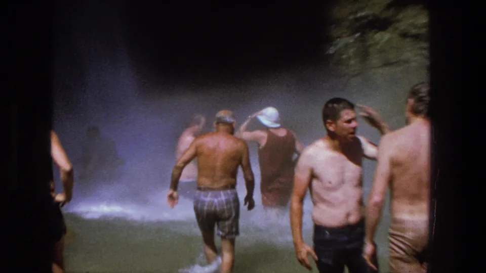 Men in swimwear stand and wade through the misty pool area beneath the waterfall in 1969 Utah footage.