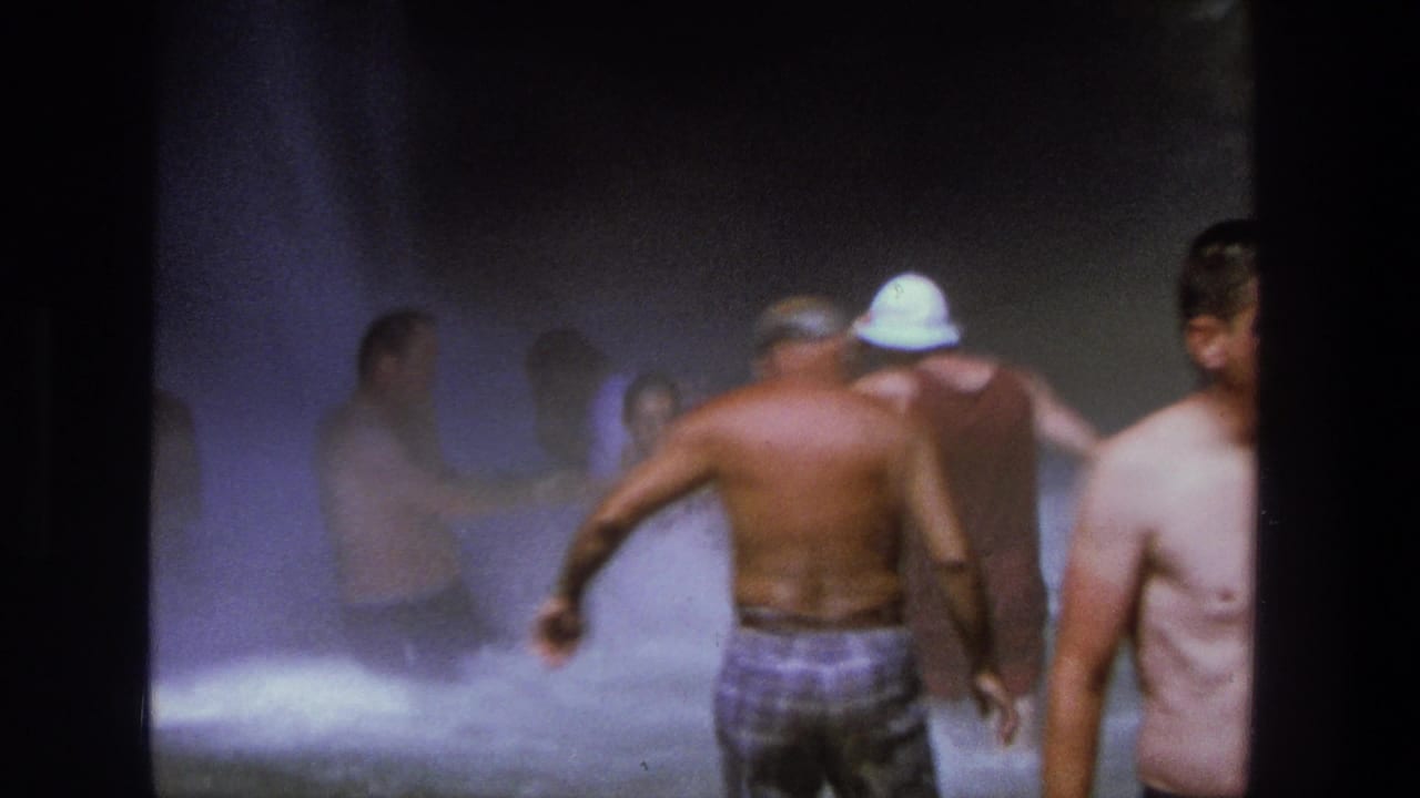 Adults wade through heavy waterfall spray, including a shirtless man in plaid swim trunks and a person in a white hat.