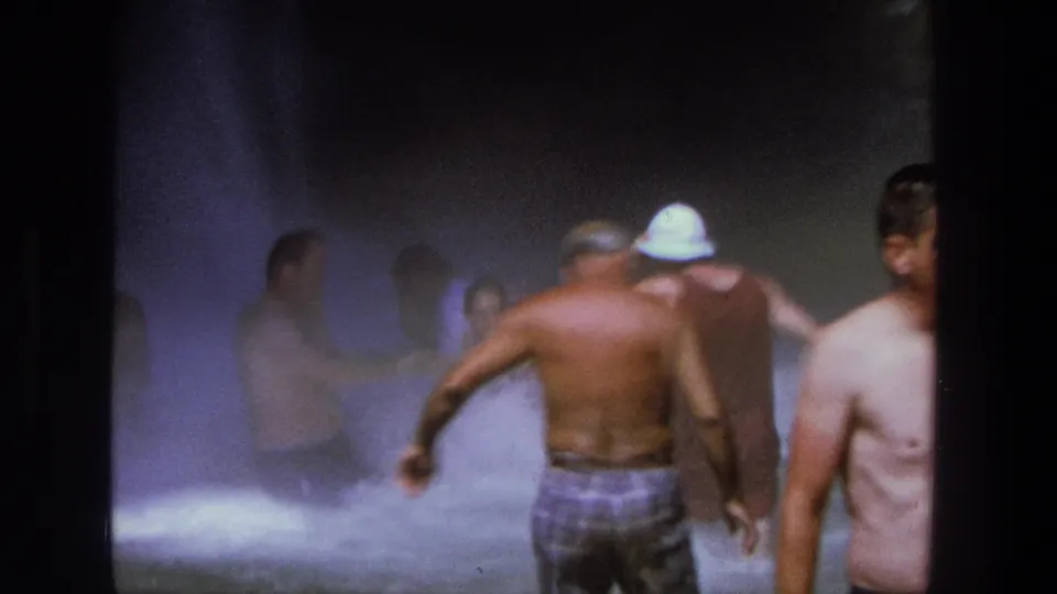 1969 Utah 8mm frame of adults moving through waterfall mist, with a shirtless man in plaid swim trunks and another person wearing a white hat.