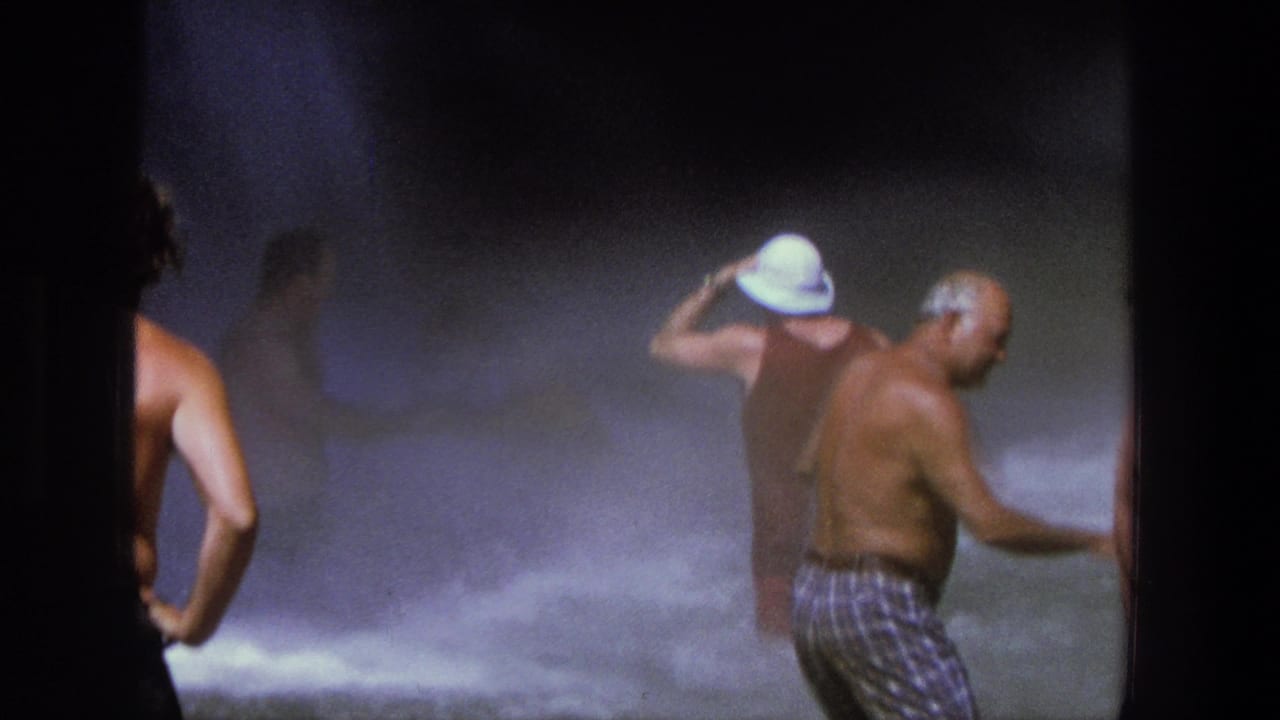 1969 Utah home-movie frame with adults in turbulent waterfall spray, including a person adjusting a white hat and a shirtless man nearby.