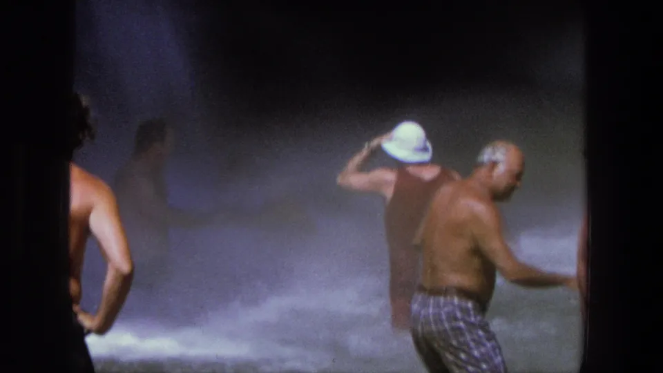 1969 Utah home-movie frame with adults in turbulent waterfall spray, including a person adjusting a white hat and a shirtless man nearby.