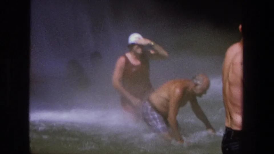 1969 Utah home-movie frame showing an adult bending in rushing water while another figure in a white hat stands amid waterfall mist.