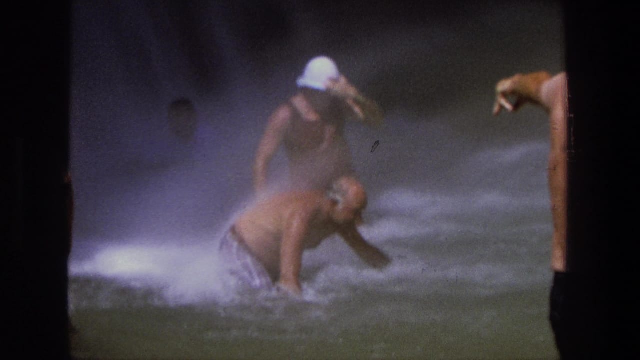 A man bends in foamy water while a swimmer in a white cap stands nearby and another arm points from shore.