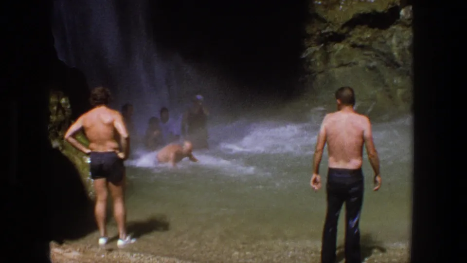 Wide 1969 Utah home-movie frame with shirtless men on shore, swimmers in a misty pool, and rocky greenery beside the waterfall.