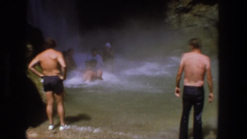 1969 Utah 8mm frame showing men in swimwear standing and wading in shallow water below a misty waterfall.
