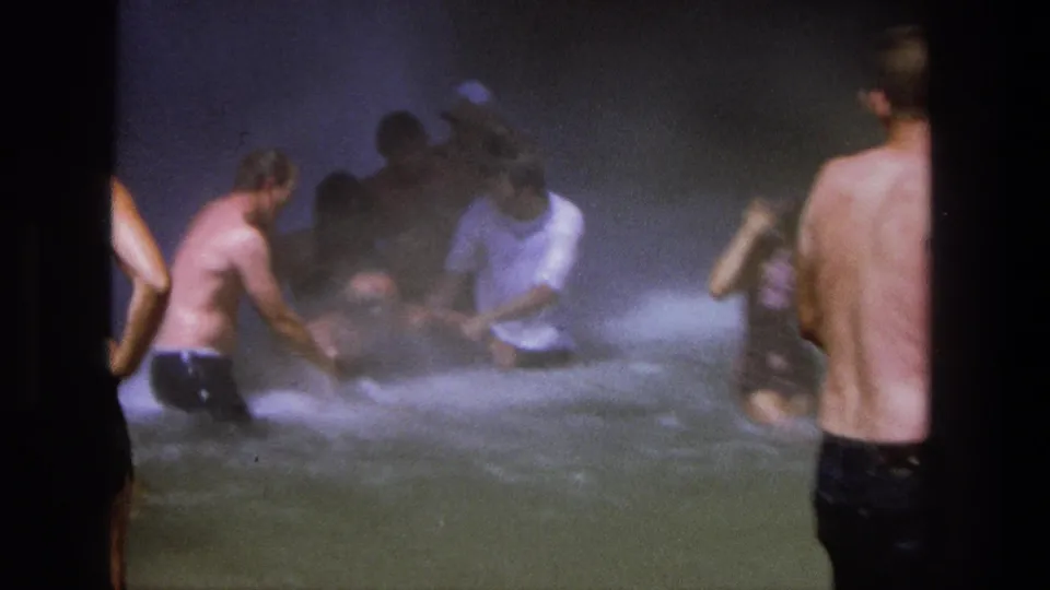 1969 Utah 8mm still of men wading and crouching in foamy water as waterfall spray softens the scene.