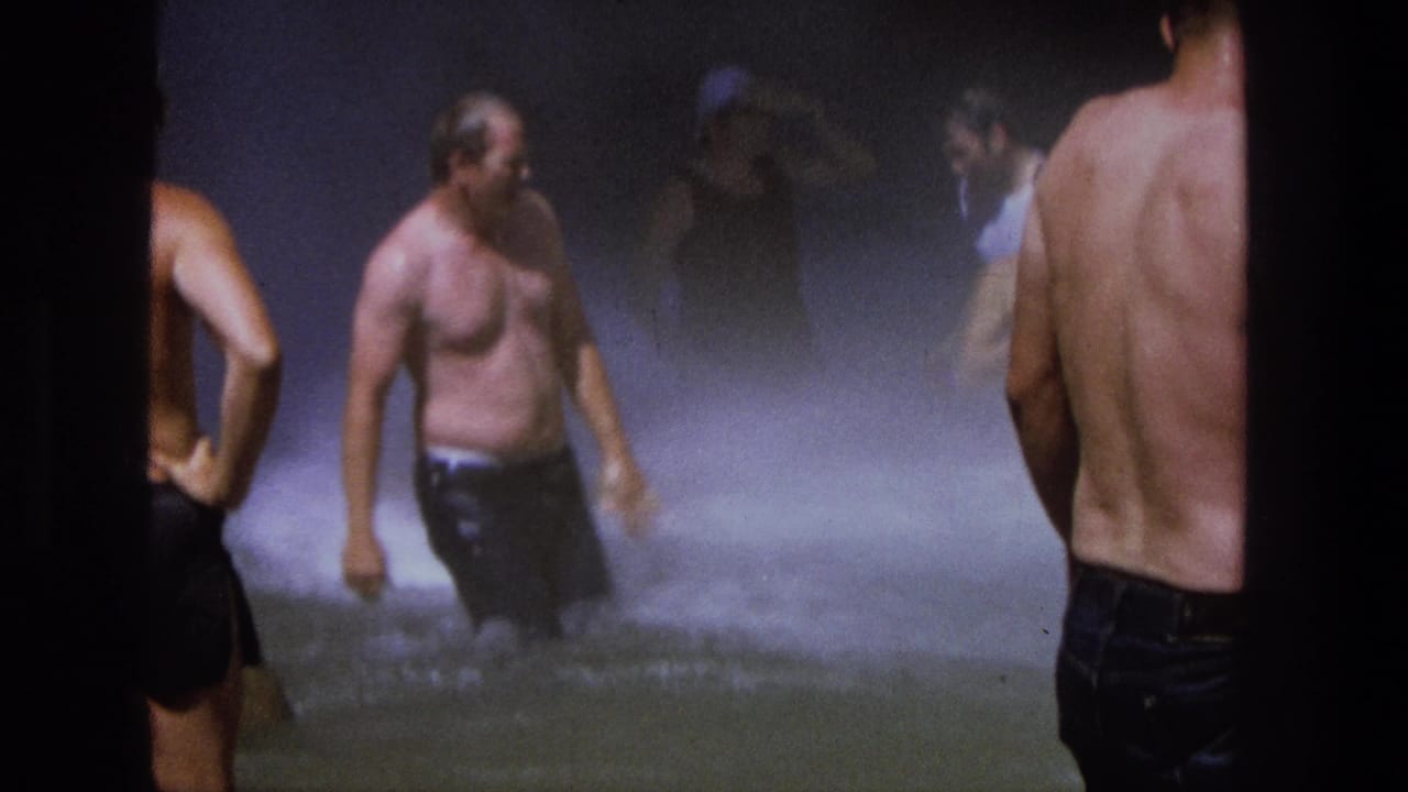 Shirtless men stand and walk through mist-filled shallow water.