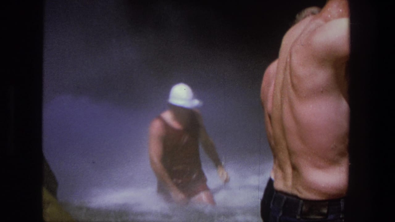 Man in a white hat walks through waterfall spray beside a shirtless observer.
