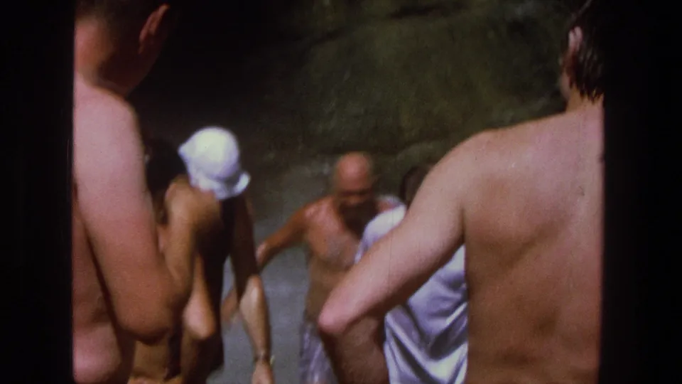 1969 Utah 8mm frame showing several men in swimwear gathered in shallow water near waterfall spray, viewed from behind onlookers.