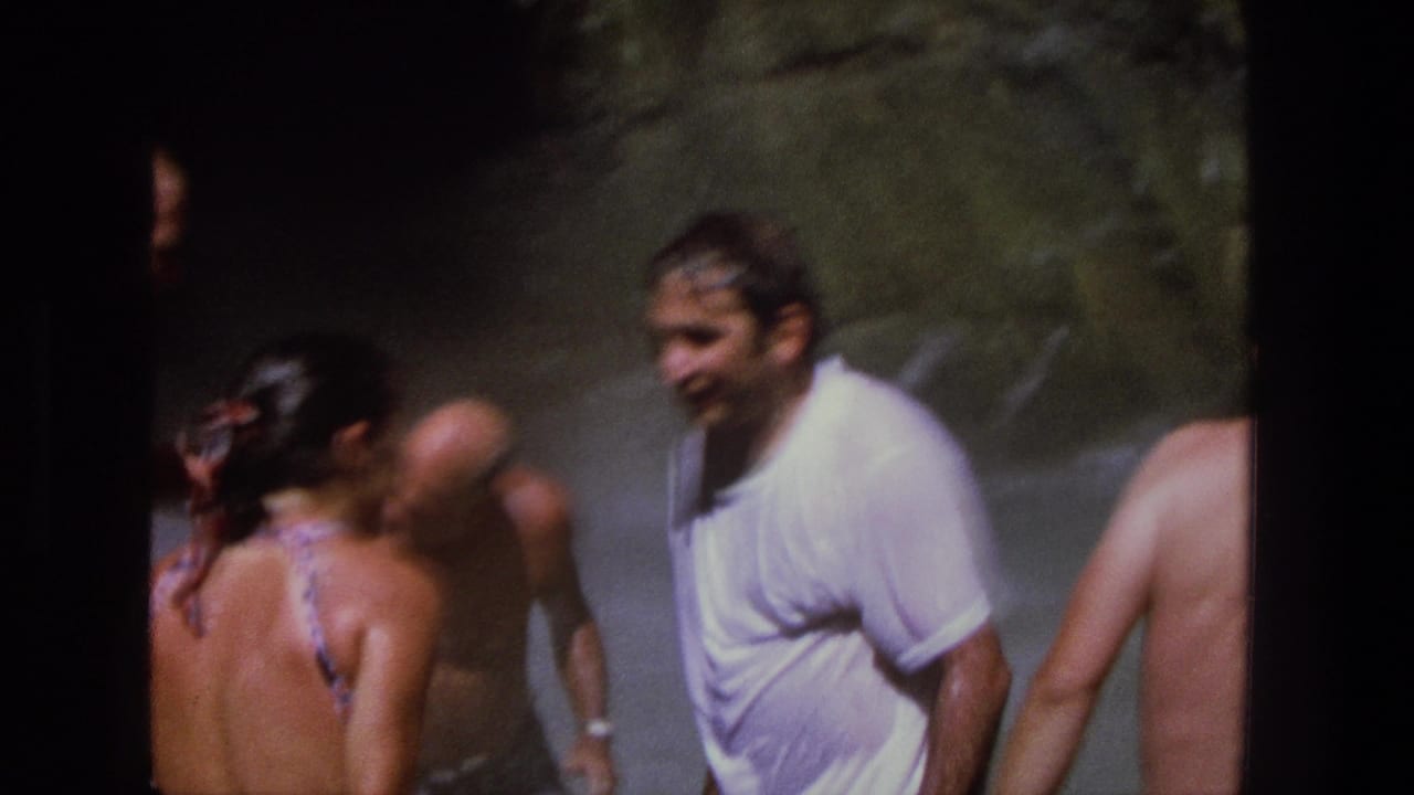 Man in a white T-shirt stands beside swimmers in misty water.