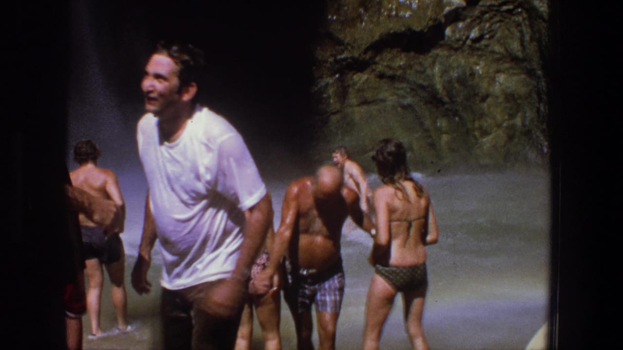 Adults in swimwear stand in waterfall spray while a man in a white T-shirt smiles in the foreground.