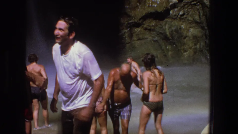 1969 Utah home-movie frame of adults wading near a rocky waterfall, with a smiling man in a white T-shirt closest to camera.