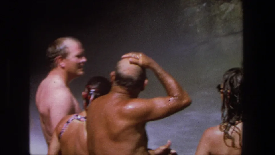 1969 Utah home-movie frame of adults in a waterfall pool, including an older man in plaid trunks lifting a hand to his head.