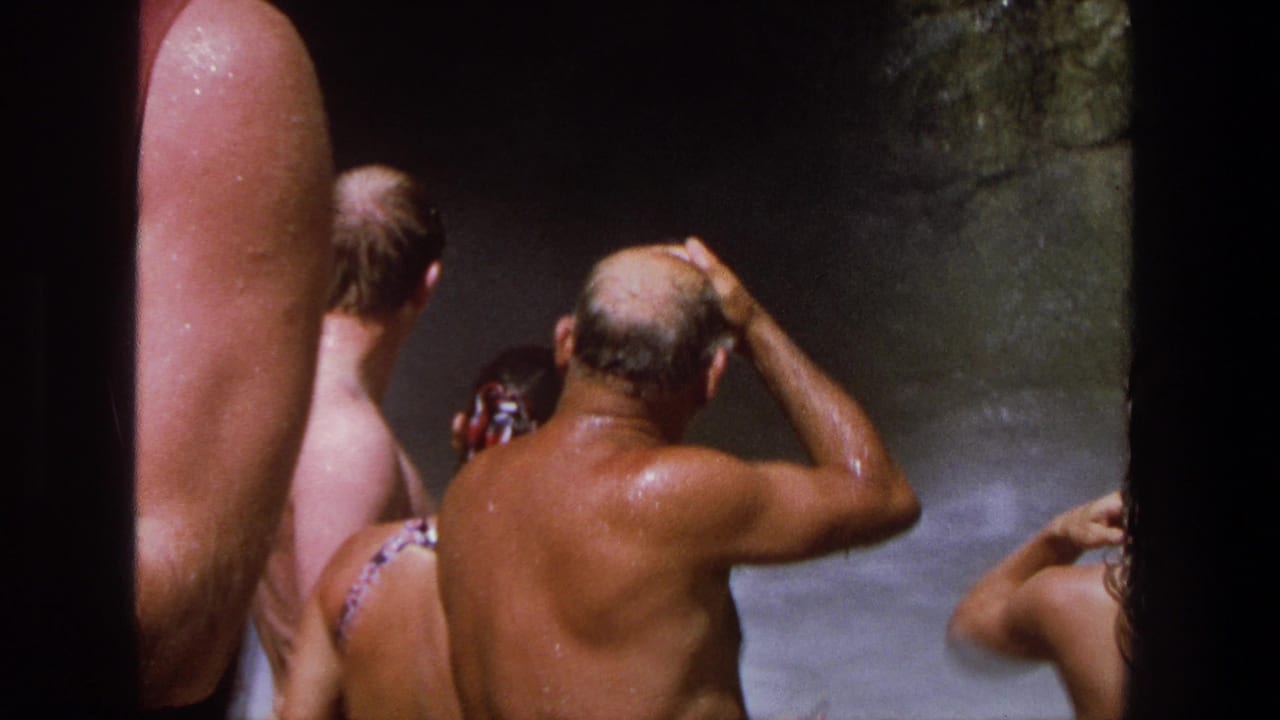 Swimmers stand near waterfall spray with wet backs and patterned swimwear visible.