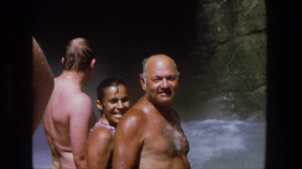 A smiling woman in a patterned swimsuit and an older man stand near the rocky watercourse in this 1969 Utah home movie.