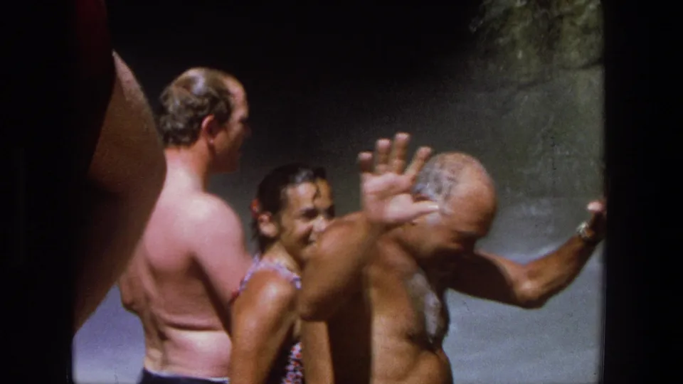 The older man lifts a hand as if waving or shielding water while other swimmers remain beside the rocky waterfall area.
